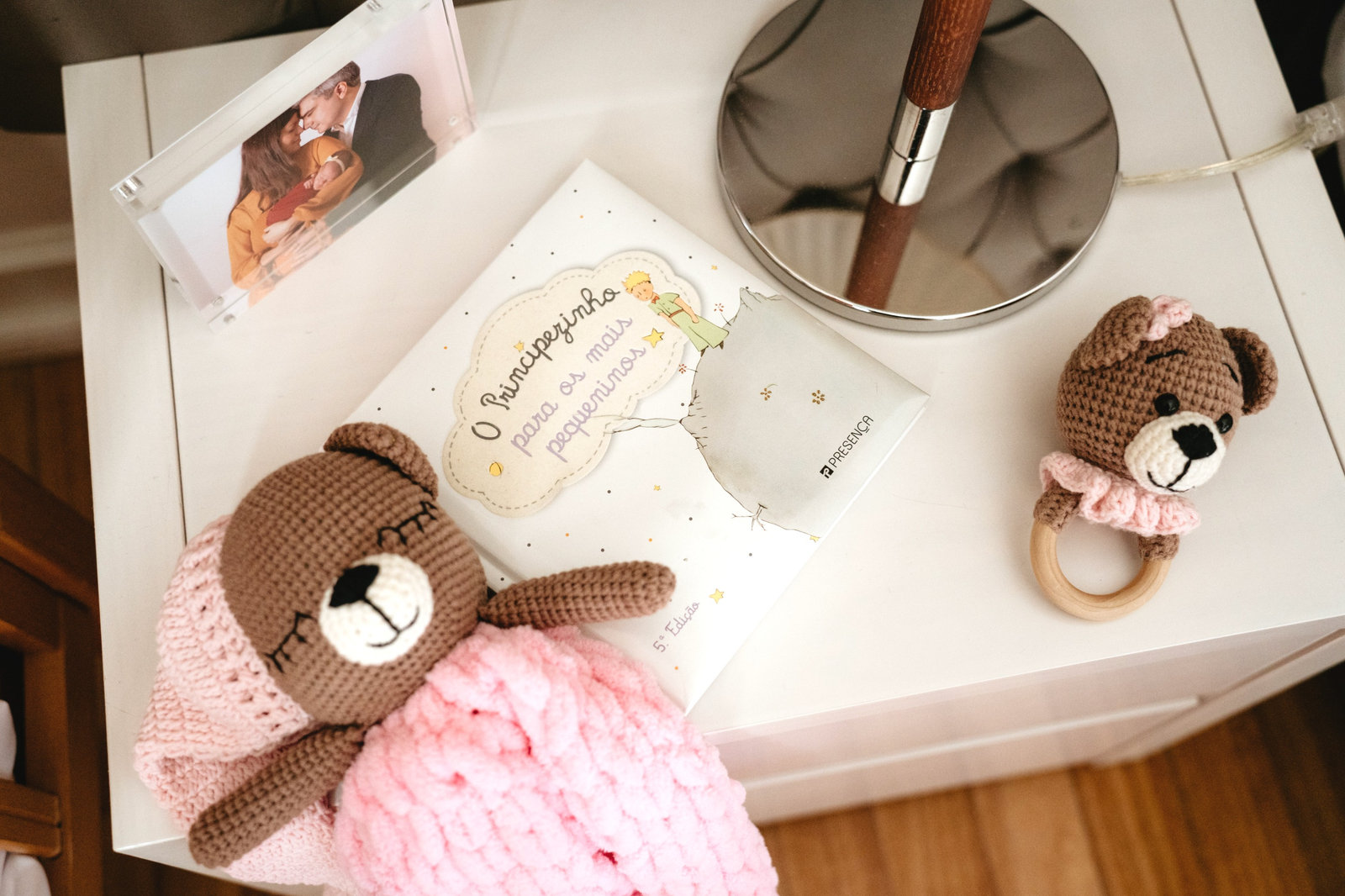 Ursinho de crochet, livro e chocalho de bebé sobre uma mesa de cabeceira, junto a uma fotografia de família