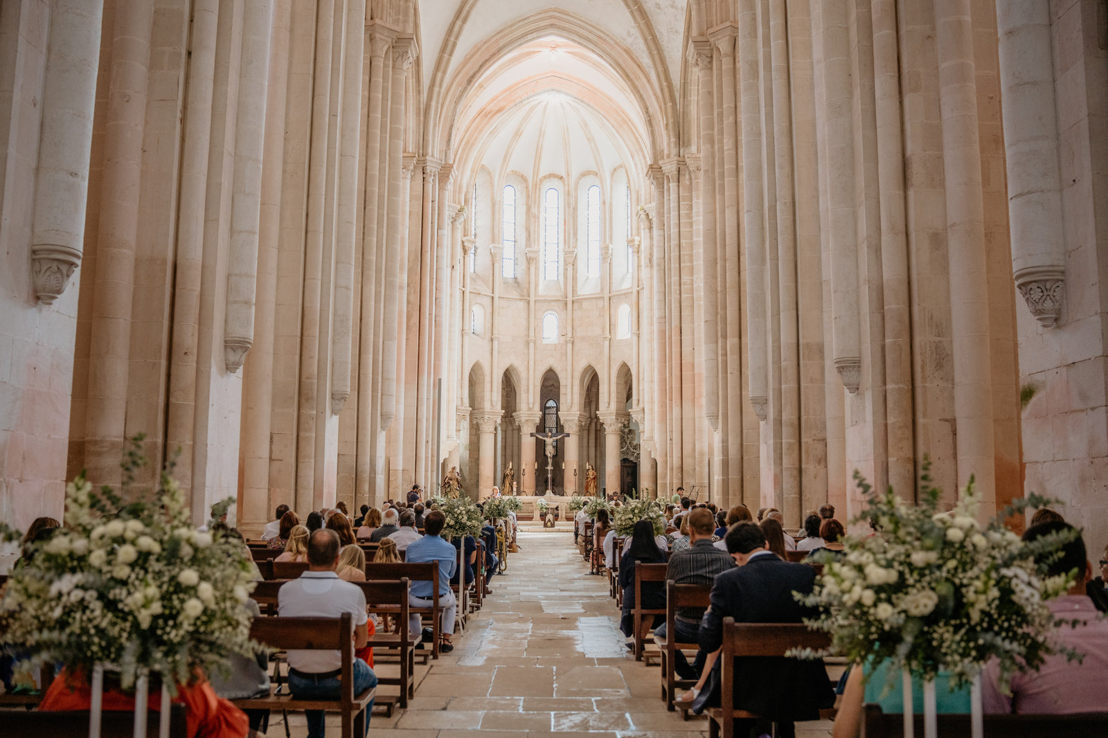 Interior do Mosteiro de Alcobaça durante uma cerimónia religiosa, com os bancos decorados com flores brancas